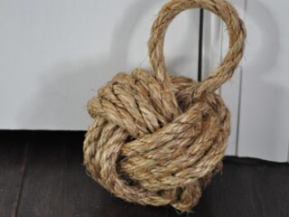 A rope knot doorstop rests against a white door on a dark wooden floor. The doorstop is made of thick, twisted rope, knotted into a ball shape with a loop handle at the top.
