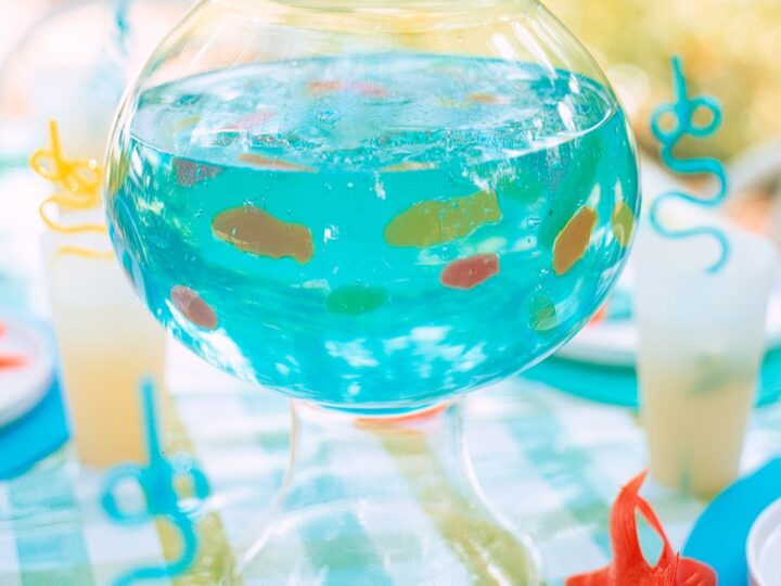 A glass bowl filled with blue liquid and gummy fish on a checkered tablecloth. Surrounding are plates, cups, and curly straws, creating a playful, aquarium-themed setting. Orange fish-shaped decorations enhance the aquatic theme.