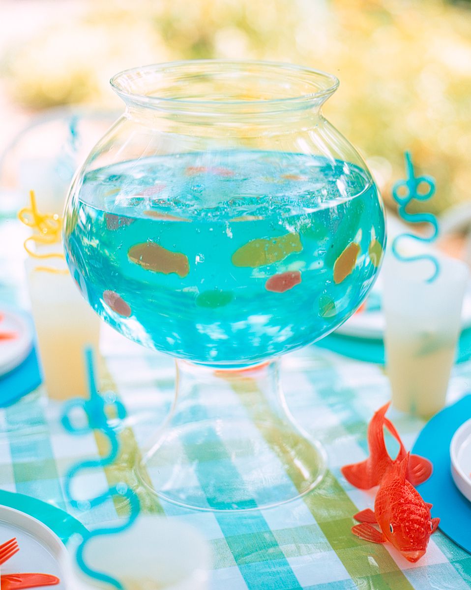 A glass bowl filled with blue liquid and gummy fish on a checkered tablecloth. Surrounding are plates, cups, and curly straws, creating a playful, aquarium-themed setting. Orange fish-shaped decorations enhance the aquatic theme.