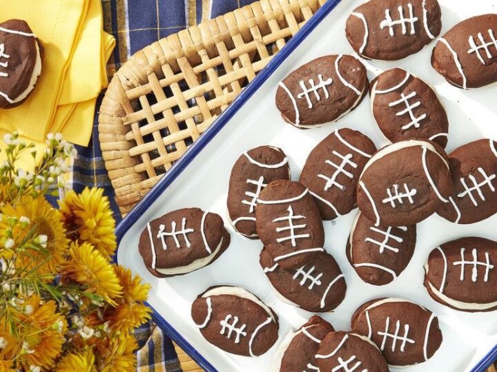 A tray of football-shaped cookies, decorated with white icing to resemble football laces, is set on a plaid tablecloth with a woven basket and a bouquet of yellow flowers nearby.