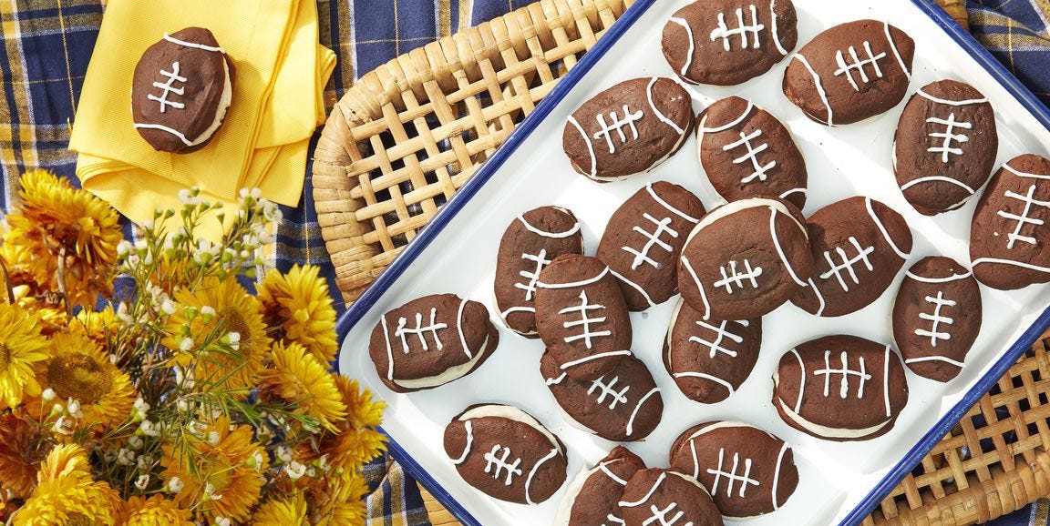 A tray of football-shaped cookies, decorated with white icing to resemble football laces, is set on a plaid tablecloth with a woven basket and a bouquet of yellow flowers nearby.