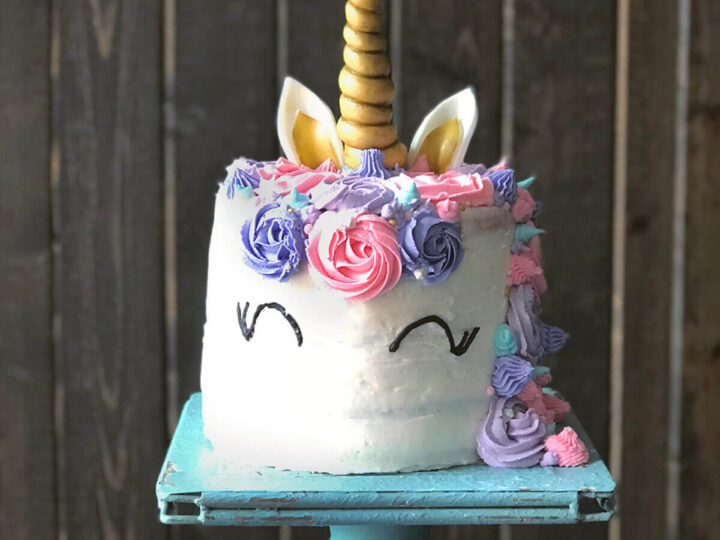 A whimsical unicorn cake with a gold horn, white frosting, and colorful pastel decorations, including roses and ears, on a teal cake stand against a wooden backdrop.