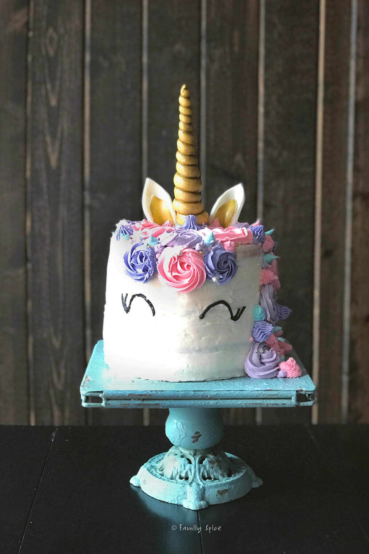 A whimsical unicorn cake with a gold horn, white frosting, and colorful pastel decorations, including roses and ears, on a teal cake stand against a wooden backdrop.