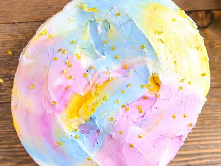 A round cake with a swirled pastel rainbow icing in shades of pink, blue, and yellow. Its decorated with small gold star sprinkles, set on a wooden surface.