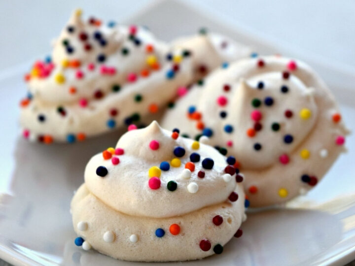 A plate of whimsical meringue cookies topped with colorful sprinkles. The text above reads Unicorn Poop Meringue Cookies. The website drugstoredivas.net is displayed at the bottom.