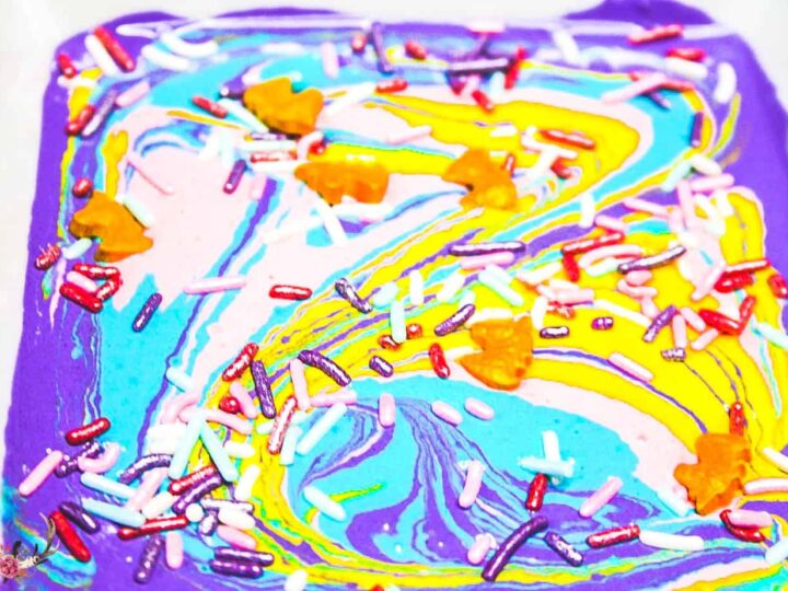 A colorful swirl of purple, blue, yellow, and pink melted candy is topped with multicolored sprinkles and small orange leaf-shaped decorations, creating a vibrant and artistic treat.