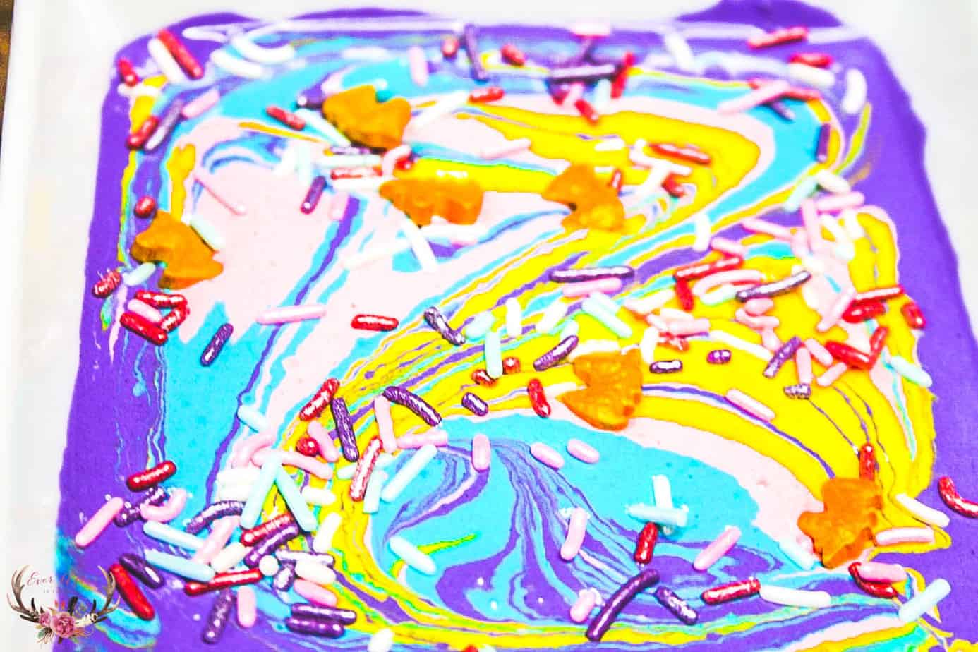 A colorful swirl of purple, blue, yellow, and pink melted candy is topped with multicolored sprinkles and small orange leaf-shaped decorations, creating a vibrant and artistic treat.