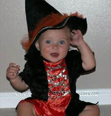 A baby wearing an orange and black witch costume with a large pointed hat is sitting on the floor. The baby is smiling and holding the brim of the hat with one hand.
