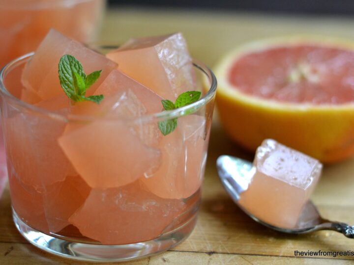 A glass filled with pink grapefruit jelly cubes, garnished with mint leaves. A half-cut grapefruit and a spoon holding a jelly cube are placed beside the glass on a wooden surface.