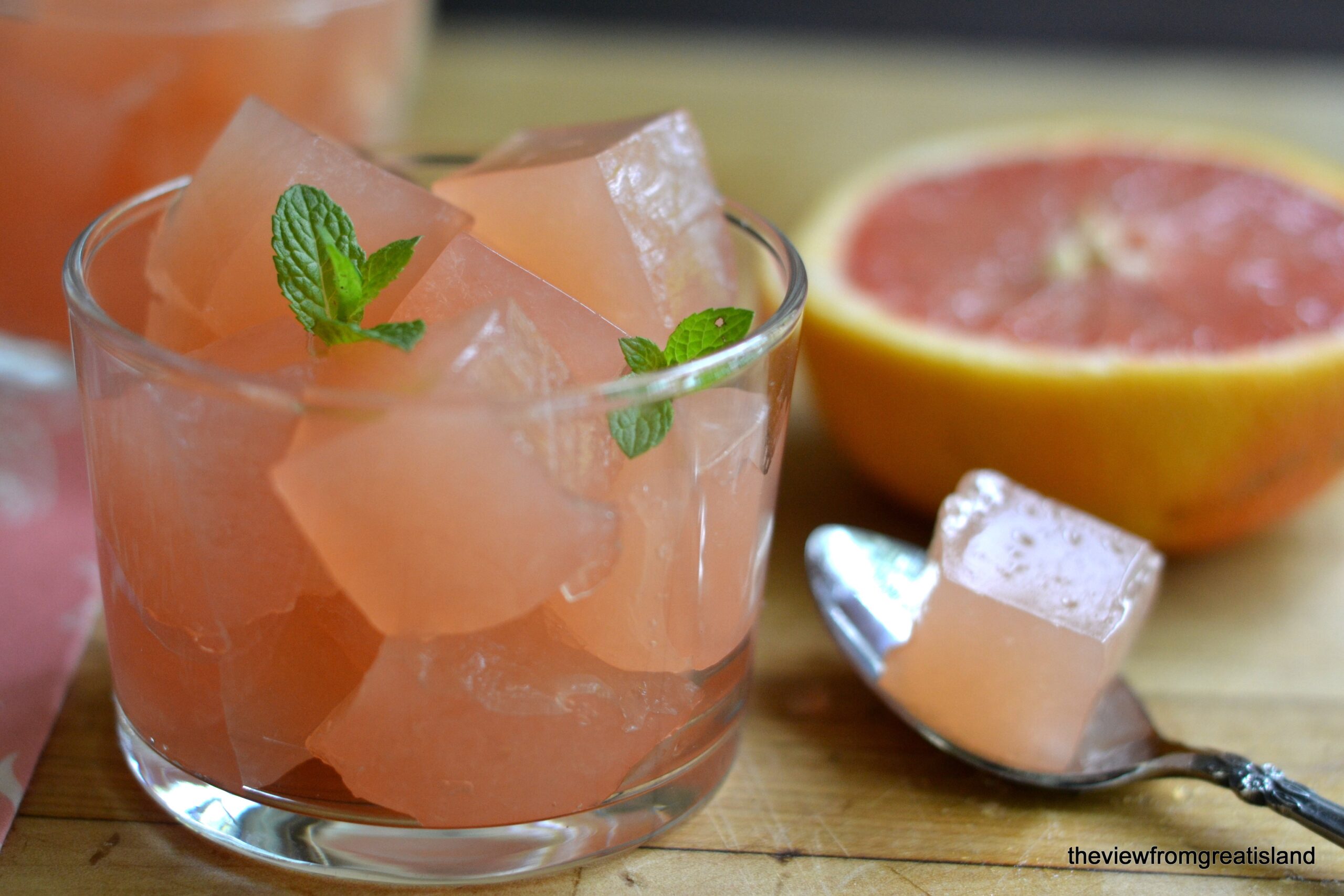 A glass filled with pink grapefruit jelly cubes, garnished with mint leaves. A half-cut grapefruit and a spoon holding a jelly cube are placed beside the glass on a wooden surface.