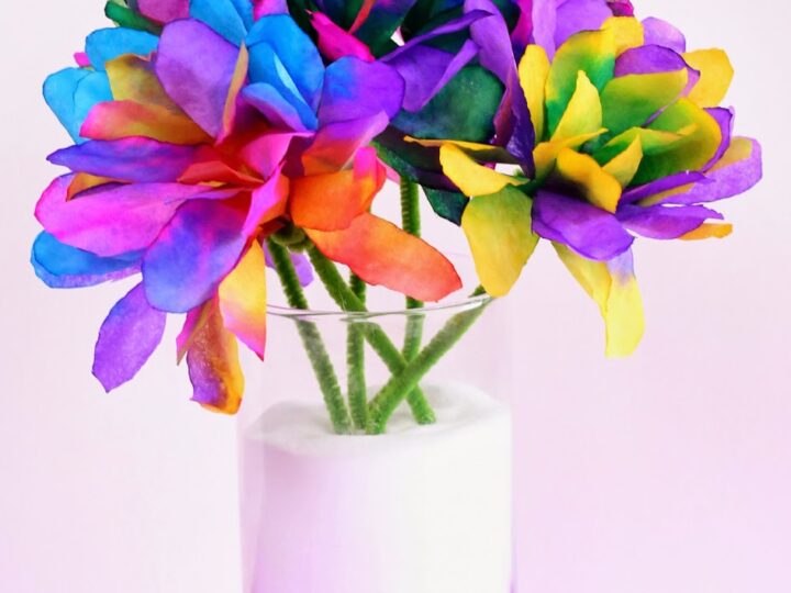 A bouquet of vibrant, multicolored flowers arranged in a clear glass vase filled with white material. The petals display a tie-dye effect with shades of blue, purple, pink, yellow, and green against a bright pink background.
