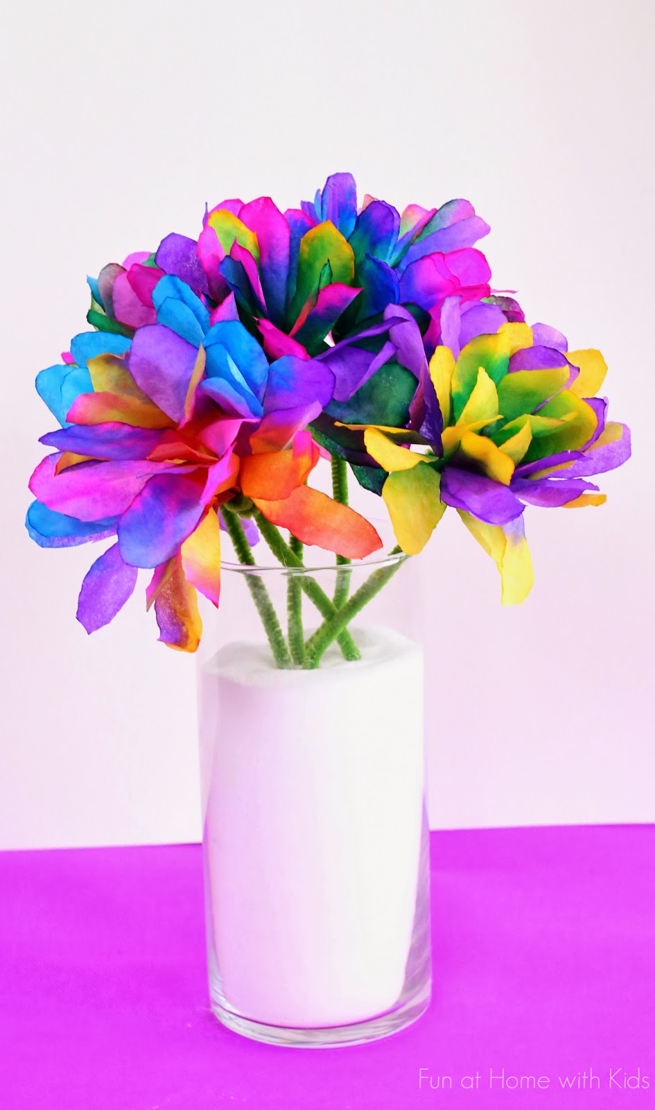 A bouquet of vibrant, multicolored flowers arranged in a clear glass vase filled with white material. The petals display a tie-dye effect with shades of blue, purple, pink, yellow, and green against a bright pink background.