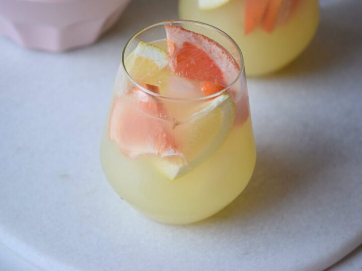 A glass of refreshing citrus punch filled with ice and slices of orange and lemon, sitting on a white round surface. Another similar glass is partially visible in the background.