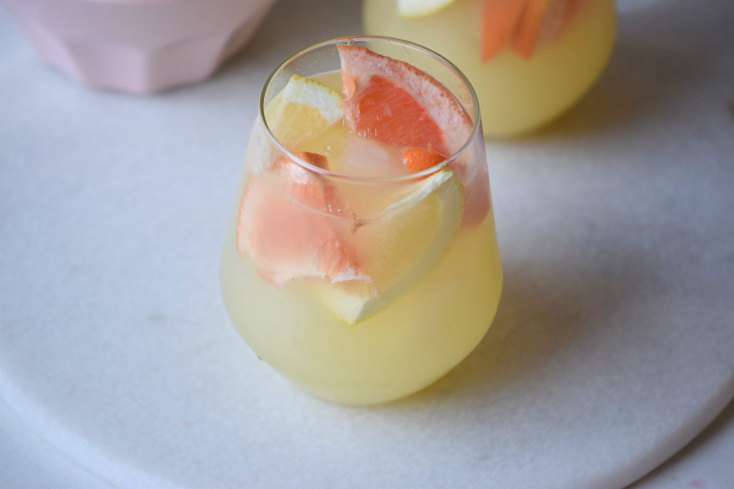 A glass of refreshing citrus punch filled with ice and slices of orange and lemon, sitting on a white round surface. Another similar glass is partially visible in the background.