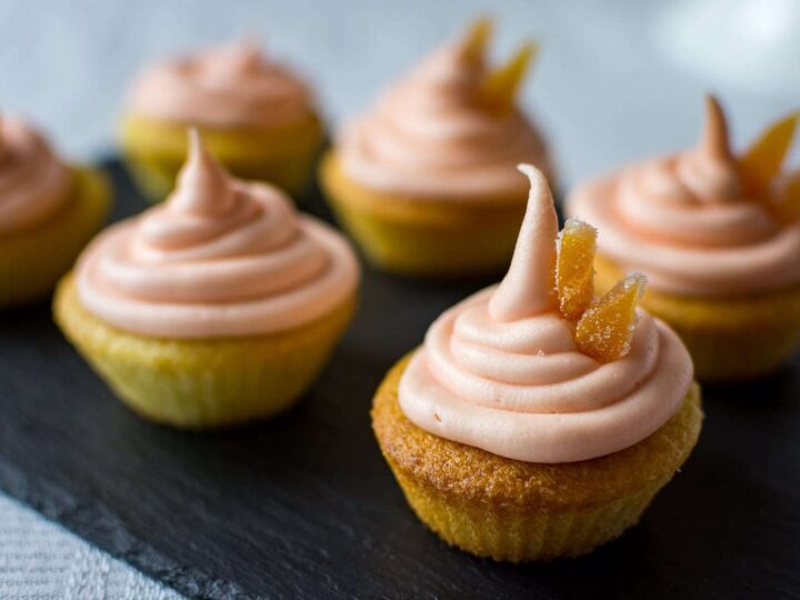 Cupcakes topped with pink frosting and candied ginger pieces are arranged on a dark slate surface. The cupcakes have a golden-brown base, and the soft frosting is swirled into peaks. The background is softly blurred.