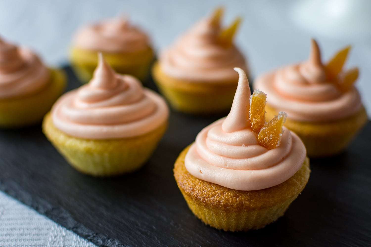 Cupcakes topped with pink frosting and candied ginger pieces are arranged on a dark slate surface. The cupcakes have a golden-brown base, and the soft frosting is swirled into peaks. The background is softly blurred.