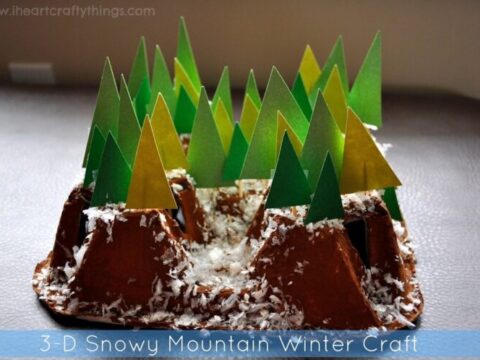 A 3D winter craft featuring brown paper mountains with green and yellow paper trees. Snow-like material is scattered on the mountains, creating a snowy scene. Text reads 3-D Snowy Mountain Winter Craft.