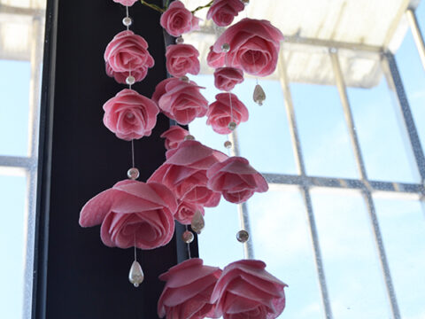 Hanging decorative piece of pink fabric roses and dangling pearl-like beads by a window, with a view of a clear blue sky and part of a building in the background.