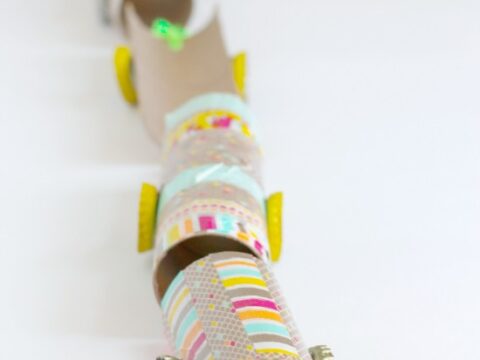 A line of colorful, handmade train cars made from cardboard rolls and bright bottle caps as wheels, decorated with patterned paper. The background is white, and the text All Aboard! is written in pink on the right.