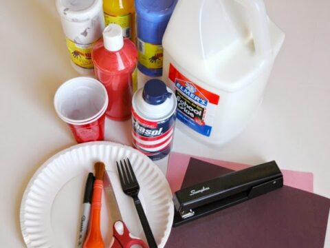 A collection of crafting supplies on a table, including bottles of paint, a gallon of glue, a stapler, scissors, a brush, a fork, a pen, a paper plate, a red cup, and a can of shaving cream.