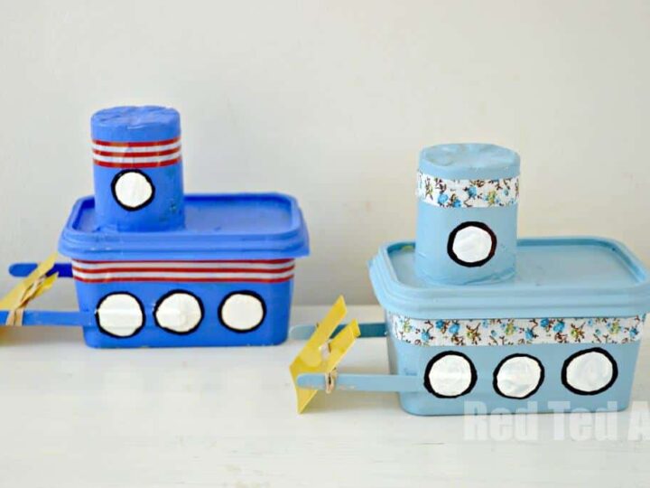 Two homemade boats made from recycled plastic containers are painted blue and light blue. Each has a cylindrical smokestack and circular windows. Yellow paddles are attached to the sides. The background is a plain white wall.
