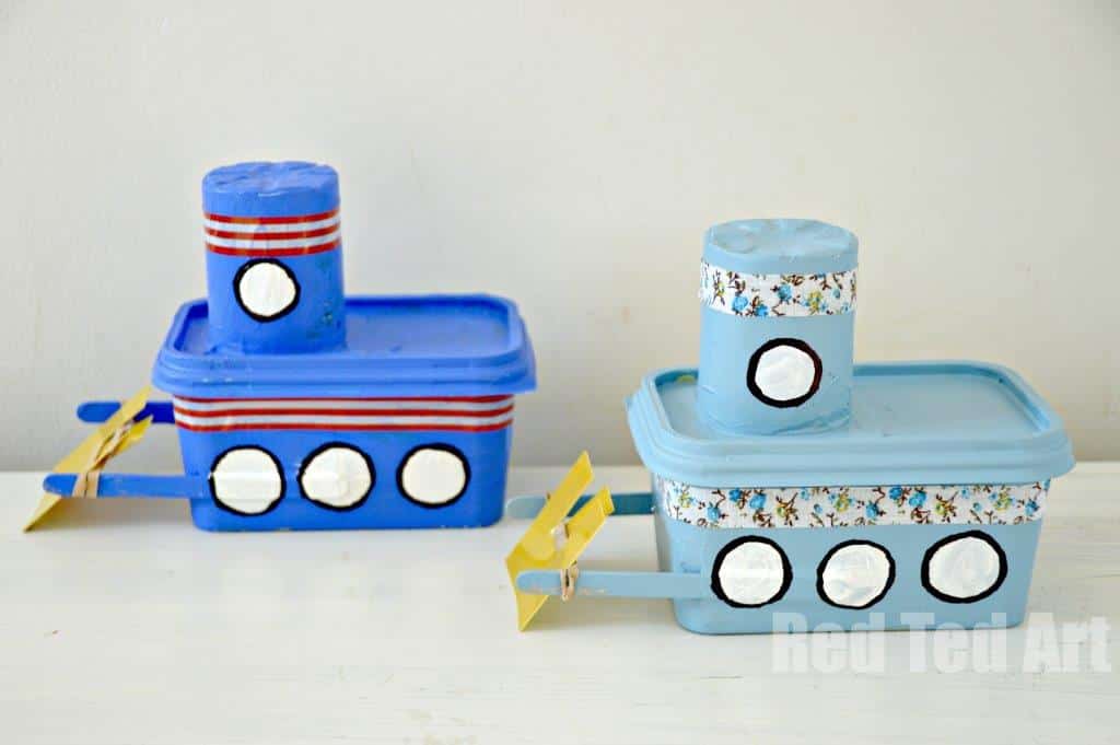 Two homemade boats made from recycled plastic containers are painted blue and light blue. Each has a cylindrical smokestack and circular windows. Yellow paddles are attached to the sides. The background is a plain white wall.