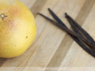 A grapefruit sits on a wooden surface with three vanilla beans placed parallel to each other in the background.