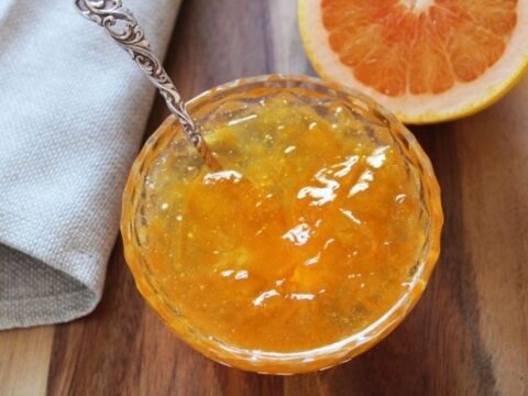 A glass dish filled with orange marmalade, placed on a wooden surface. A silver spoon rests inside the dish. In the background, theres a beige napkin and a halved orange showing its juicy interior.