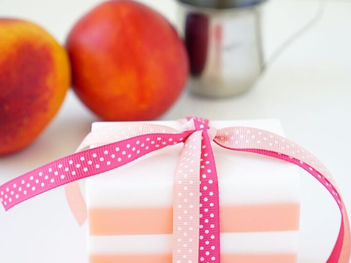 A decorative soap with pink and white stripes is tied with a pink ribbon. In the background, there are two peaches and a small metal pitcher. The scene has a clean, bright appearance.