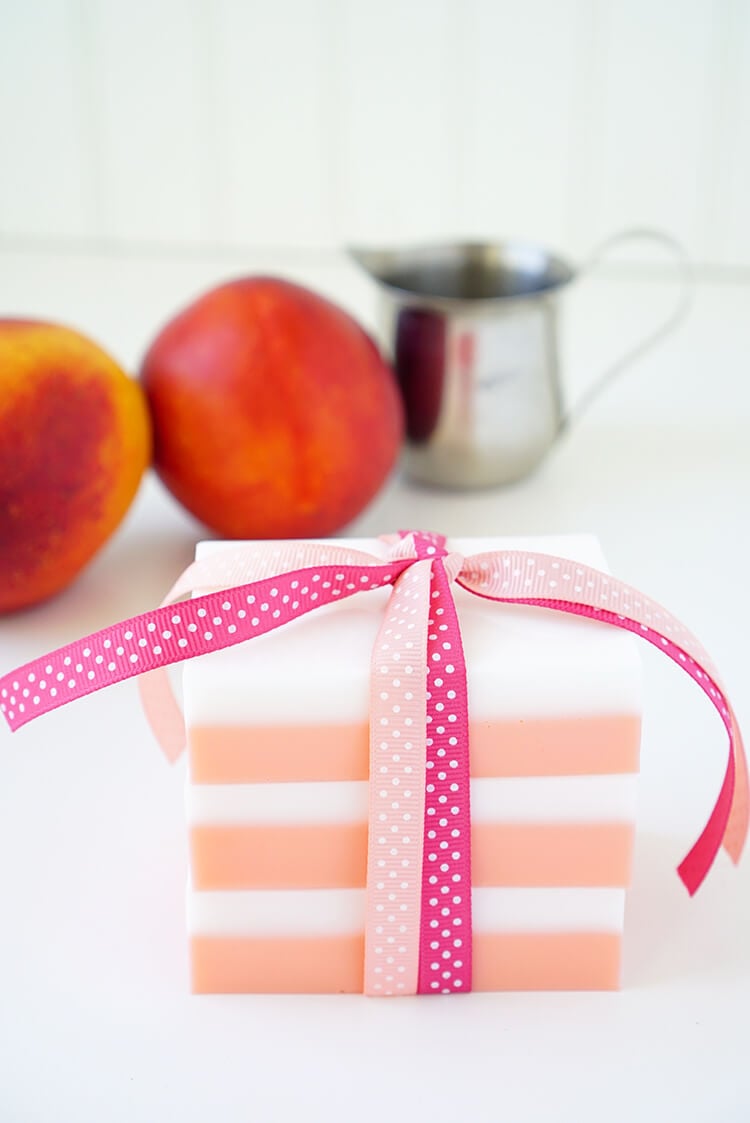 A decorative soap with pink and white stripes is tied with a pink ribbon. In the background, there are two peaches and a small metal pitcher. The scene has a clean, bright appearance.