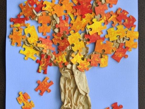 A tree crafted from brown crumpled paper for the trunk and branches, with puzzle pieces in various shades of orange, red, and yellow forming the leaves, set against a blue background with green grass at the base.