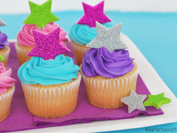 Cupcakes topped with swirls of colorful icing in pink, blue, and purple are adorned with glittery star toppers in silver, pink, green, and purple. They are arranged on a purple napkin over a white plate against a blue background.