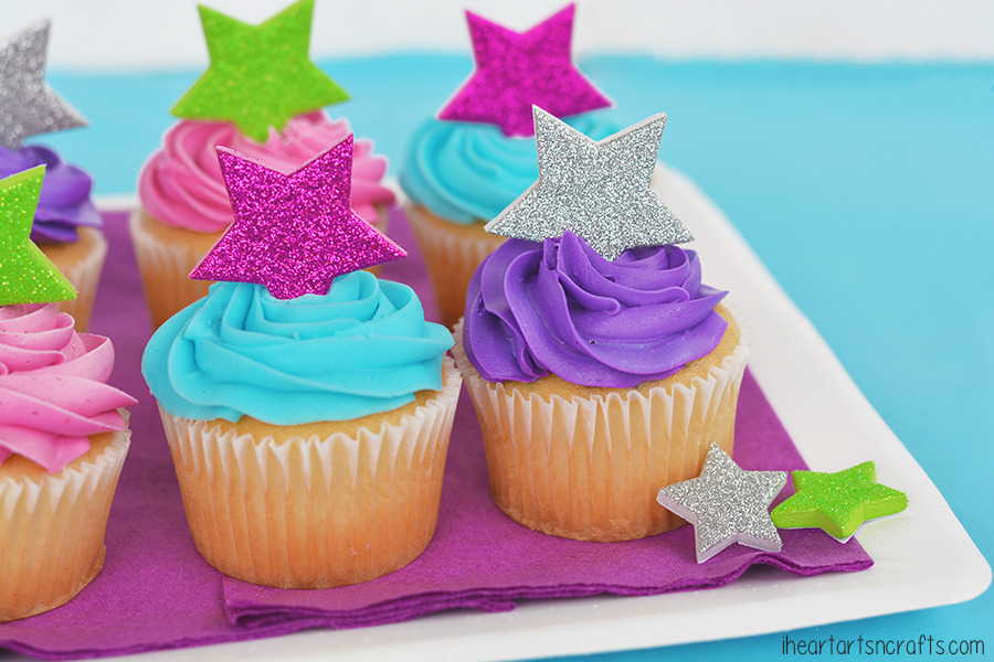 Cupcakes topped with swirls of colorful icing in pink, blue, and purple are adorned with glittery star toppers in silver, pink, green, and purple. They are arranged on a purple napkin over a white plate against a blue background.