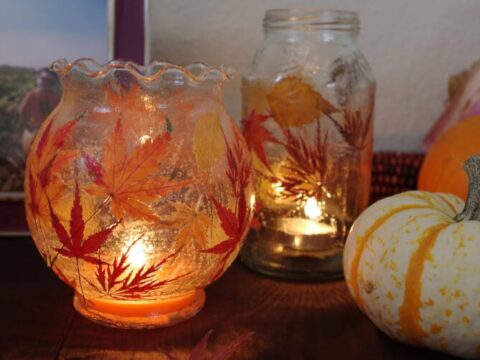 Glass jars decorated with autumn leaves hold lit tealights, casting a warm glow. A small orange and white pumpkin sits nearby, and a framed photo is partially visible in the background, creating a cozy autumn atmosphere.