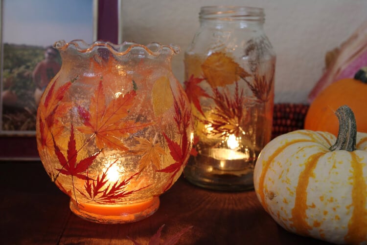 Glass jars decorated with autumn leaves hold lit tealights, casting a warm glow. A small orange and white pumpkin sits nearby, and a framed photo is partially visible in the background, creating a cozy autumn atmosphere.