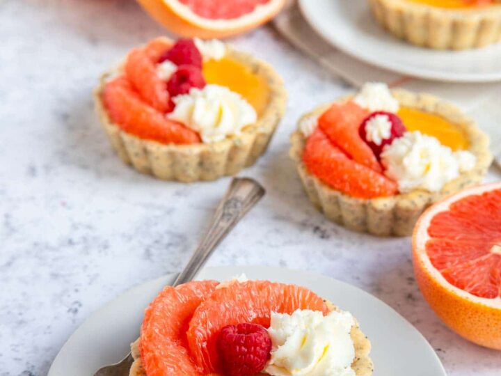 A plate with a small tart topped with grapefruit slices, raspberries, and cream. More tarts and halved grapefruits are in the background on a light marble surface. A fork rests on the plate next to the tart.