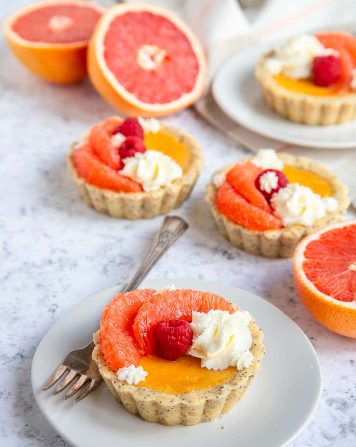 A plate with a small tart topped with grapefruit slices, raspberries, and cream. More tarts and halved grapefruits are in the background on a light marble surface. A fork rests on the plate next to the tart.