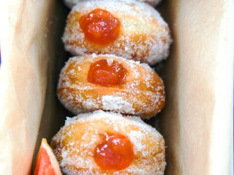 A row of sugar-coated doughnuts filled with red jelly, placed in a beige paper-lined box. Two grapefruit slices are positioned at opposite corners of the box.