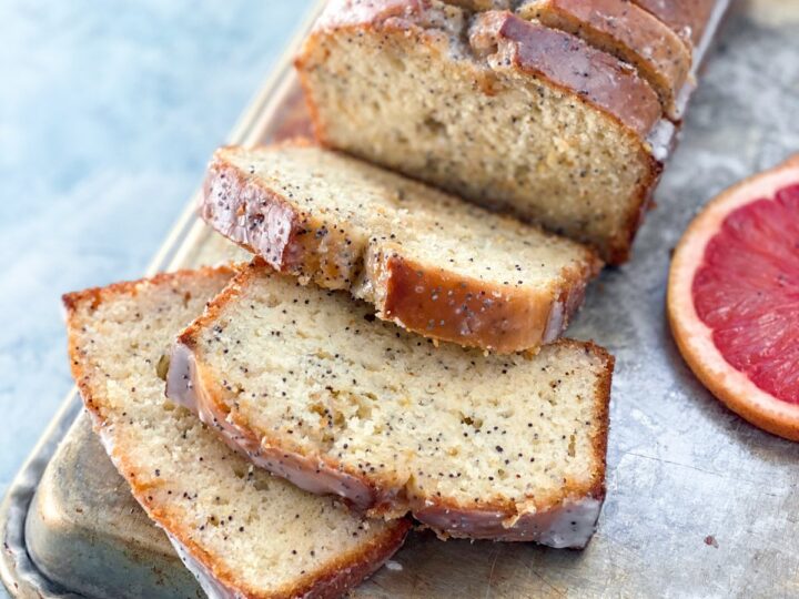 A loaf of poppy seed bread with a glazed top, sliced on a baking sheet. A few slices are laid out in front, revealing the moist interior. A half slice of grapefruit is visible next to the bread.