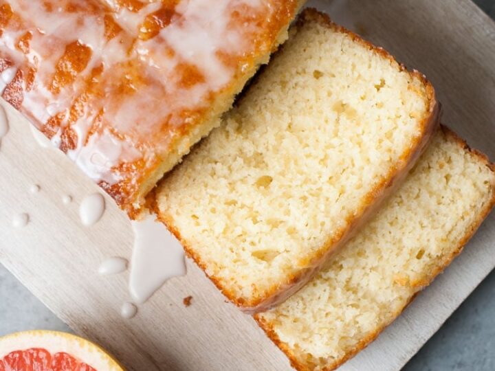 Top view of a glazed loaf cake with two slices cut, showing a moist and fluffy interior. The cake is on a wooden board with a half grapefruit nearby on a gray surface.