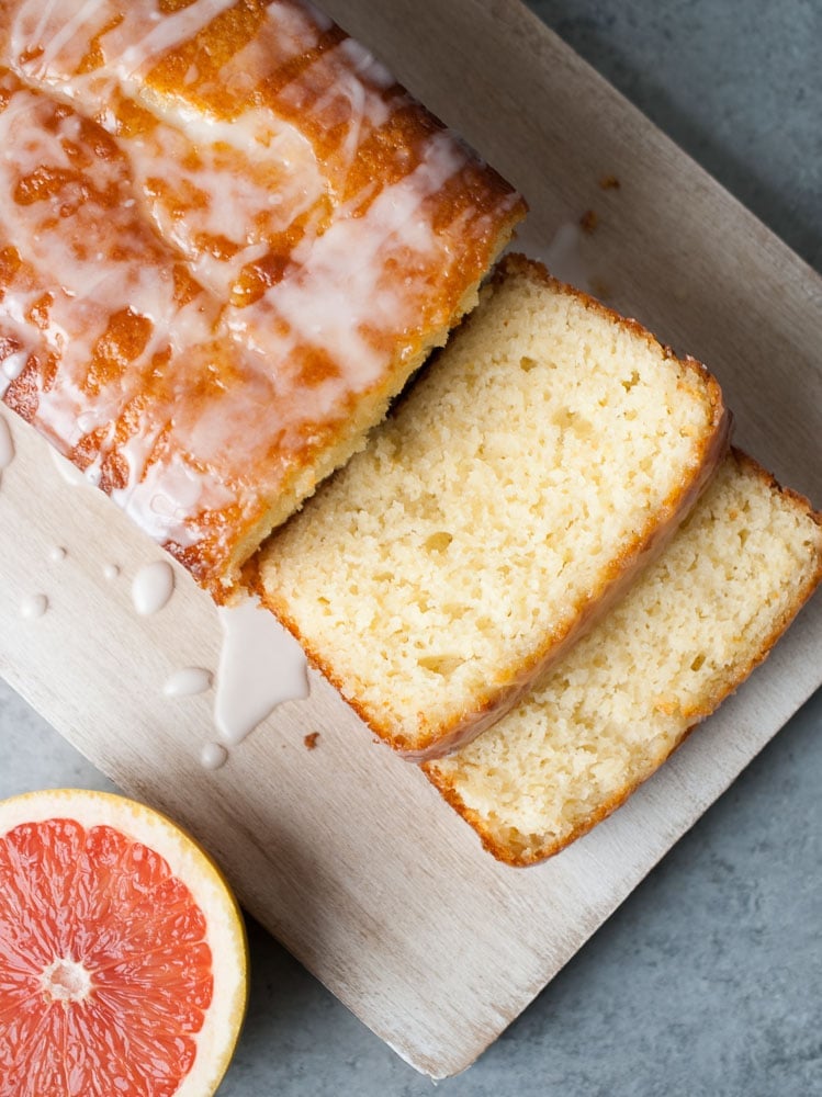 Top view of a glazed loaf cake with two slices cut, showing a moist and fluffy interior. The cake is on a wooden board with a half grapefruit nearby on a gray surface.