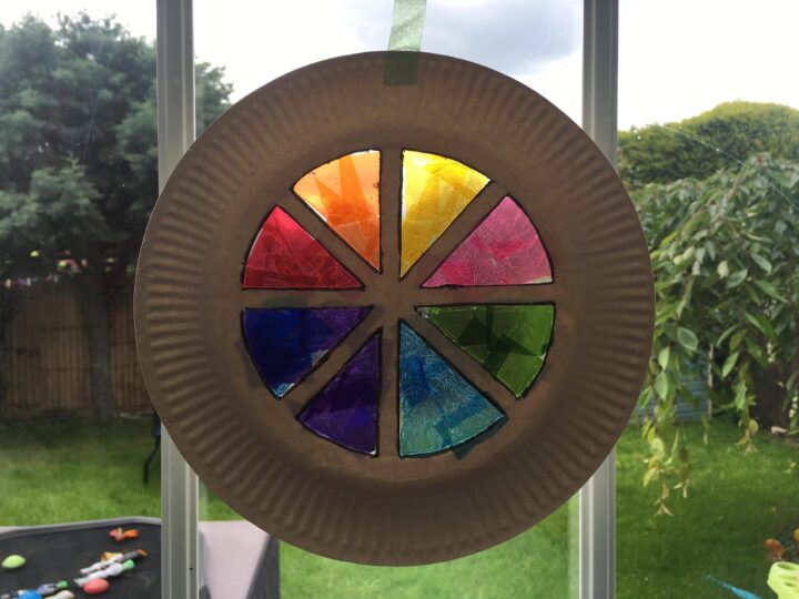 A paper plate with colorful stained glass segments hangs in a window. The colors include red, orange, yellow, green, blue, and purple, arranged in a circular pattern. A garden is visible in the background.
