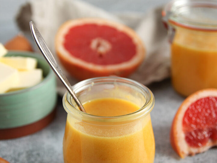 A small glass jar of grapefruit curd with a spoon inside it. Halved grapefruits and more curd in a similar jar are in the background. Butter is placed in a green bowl nearby, all set on a gray surface.