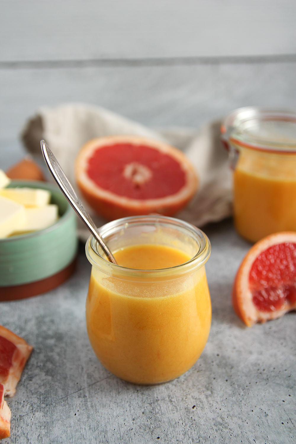 A small glass jar of grapefruit curd with a spoon inside it. Halved grapefruits and more curd in a similar jar are in the background. Butter is placed in a green bowl nearby, all set on a gray surface.
