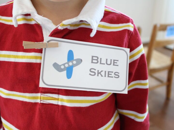 A child wearing a red and yellow striped shirt with a name tag clipped on. The tag displays an airplane icon and the text Blue Skies. A chair is blurred in the background.