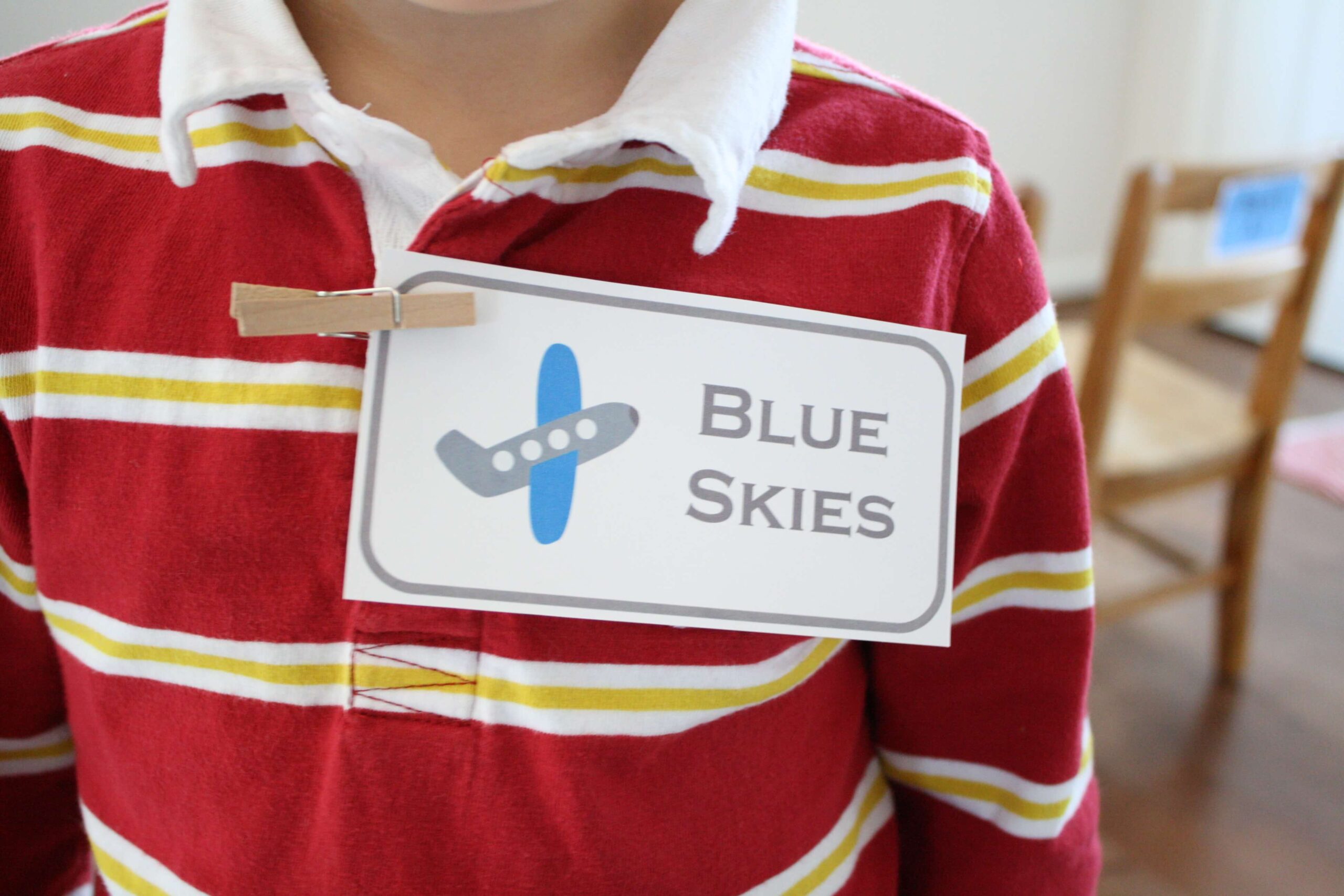 A child wearing a red and yellow striped shirt with a name tag clipped on. The tag displays an airplane icon and the text Blue Skies. A chair is blurred in the background.