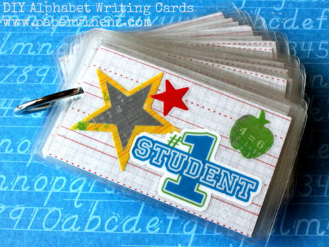 A stack of DIY alphabet writing cards held together with a metal ring on a blue background. The top card features a large gold star, a small red star, a green apple, and the text #1 Student. The website www.paperwings.com is visible at the top.
