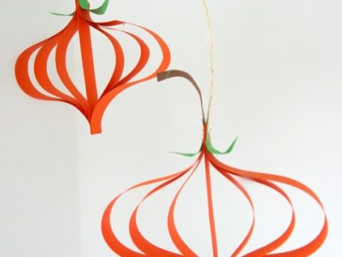 Two orange paper pumpkins hang as decorations against a white wall. They are suspended by strings and have green leaf accents and brown stems.