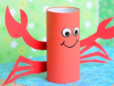 A cute crab craft made from a red toilet paper roll with googly eyes and a smile. It has red paper claws and legs. The background is a green floral pattern with a blue sea-themed surface.