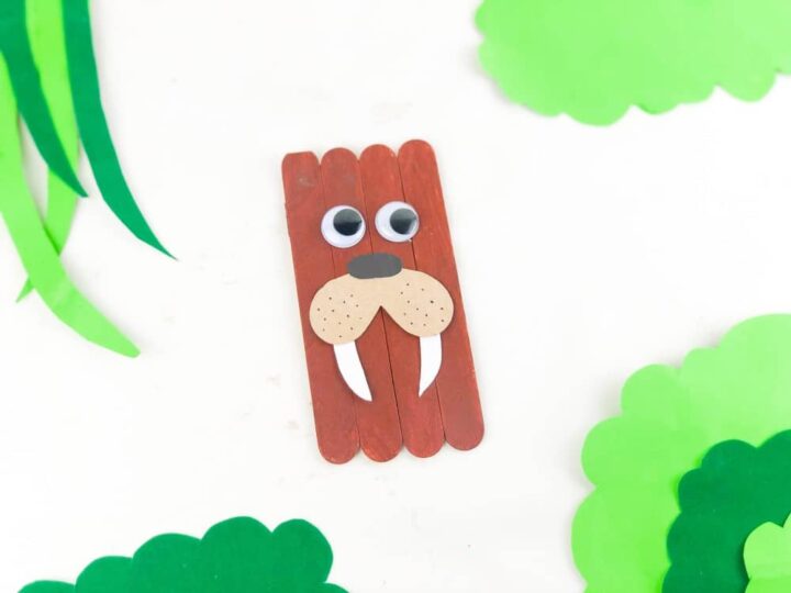 Craft depicts a walrus, made from brown popsicle sticks arranged vertically. It features large googly eyes, white tusks, and a round nose. Green paper cutouts surround it, resembling leaves or foliage.
